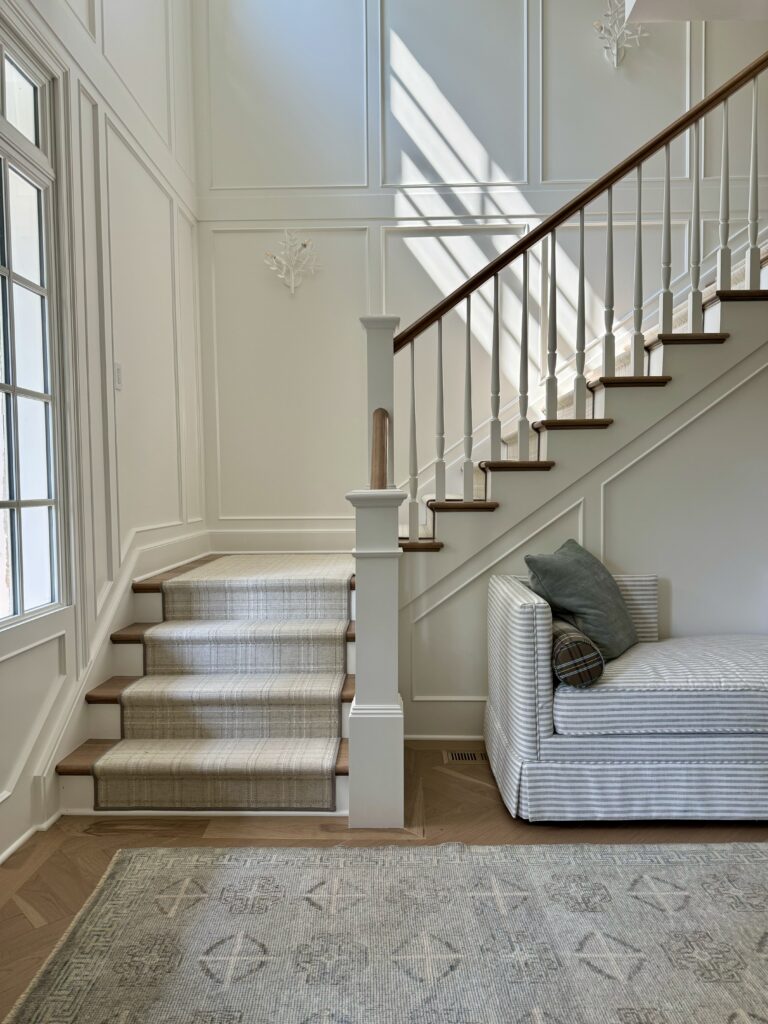 Completed foyer transformation with light and airy decor, white walls, detailed moldings, a patterned stair runner, and a cozy sofa. Perfect for modern interior design inspiration.